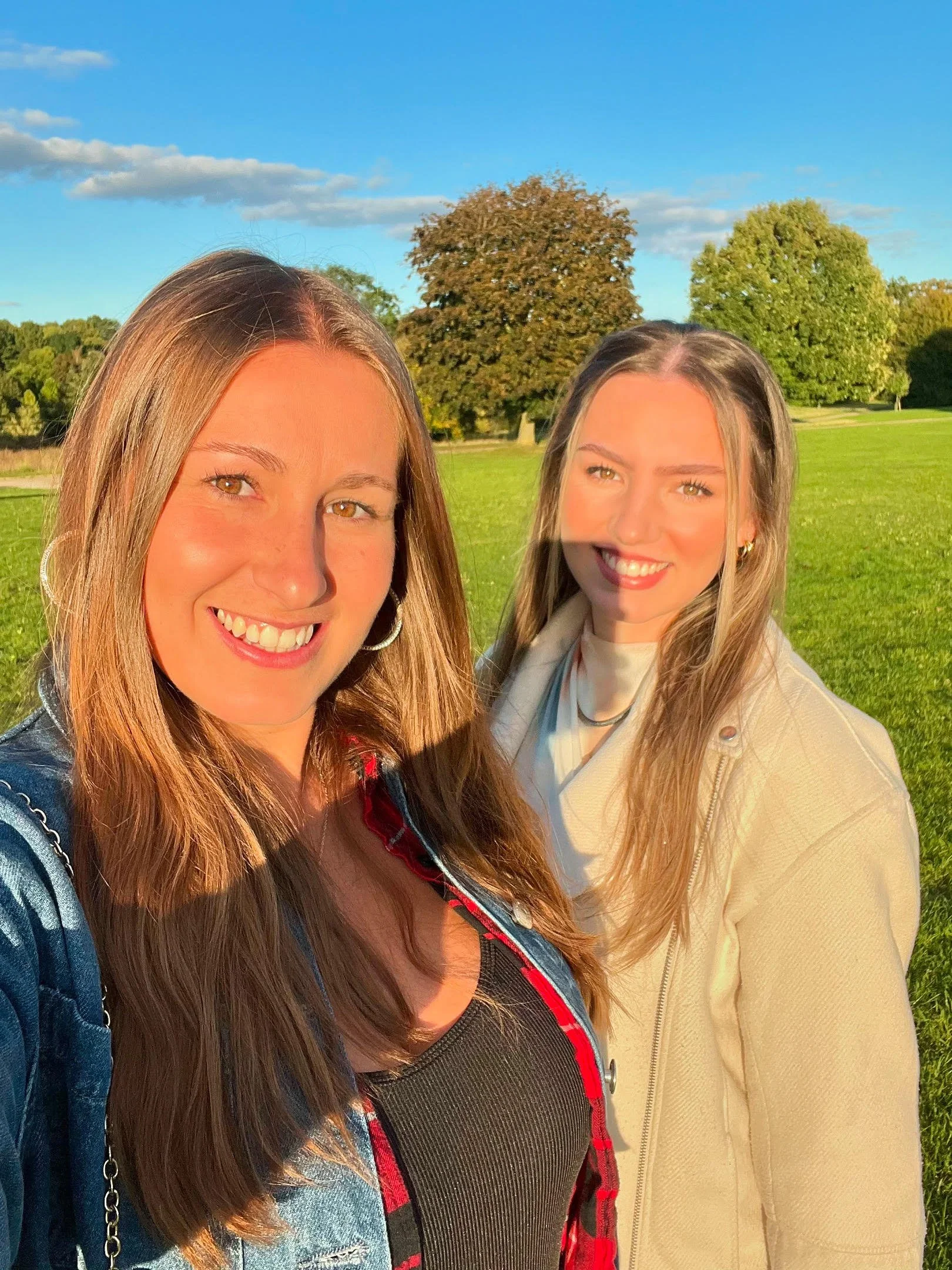 Two smiling women standing outdoors on a sunny day with green grass and large trees in the background.