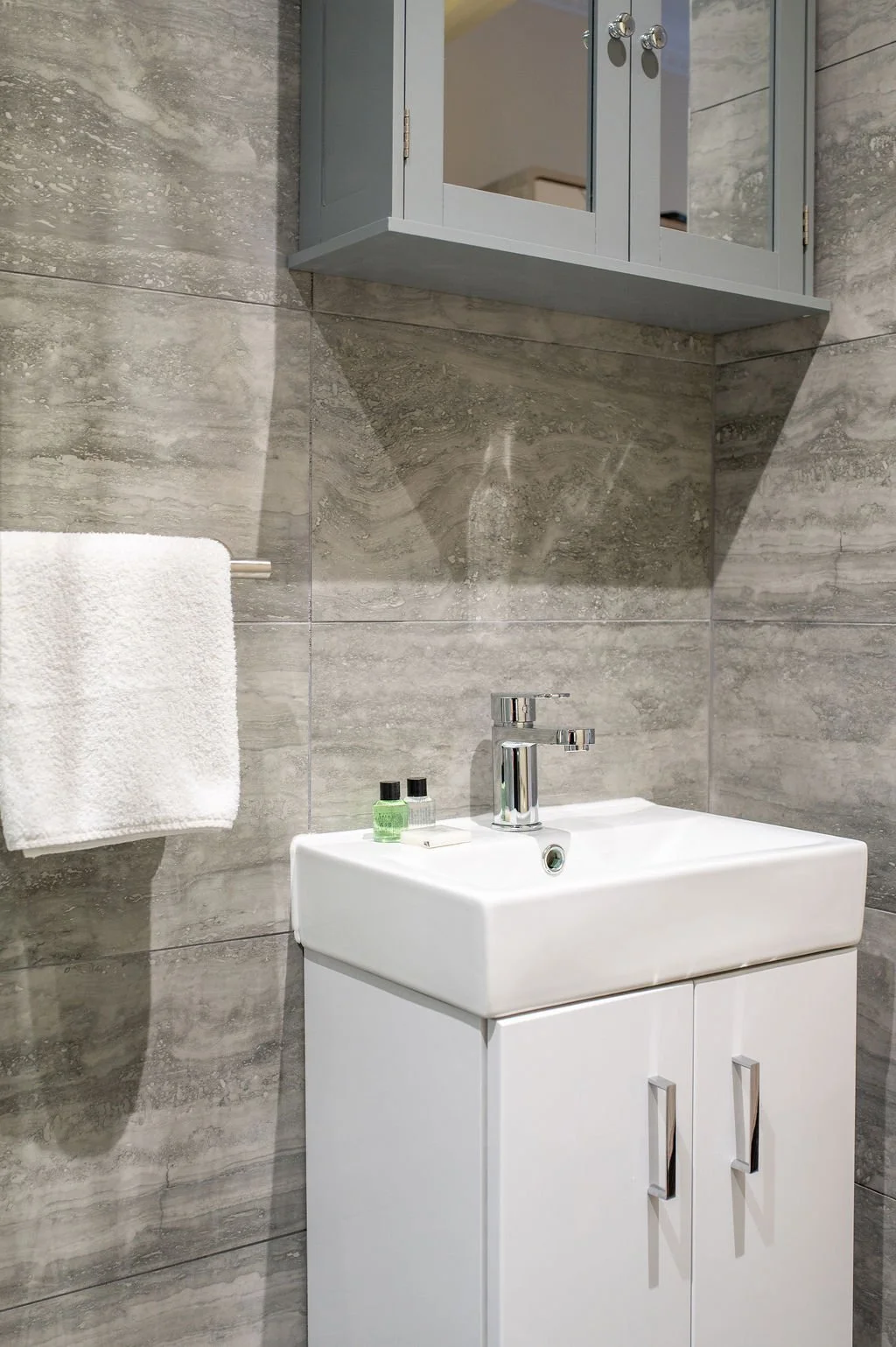 A modern bathroom with a white sink cabinet, a grey wall-mounted cabinet with mirrored doors, and a towel hanging on a metal bar.