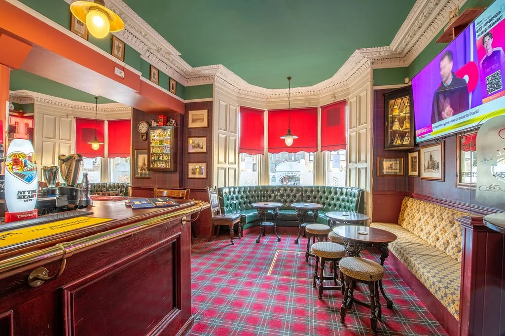 A cozy pub interior with a wooden bar counter, green upholstered seating, small tables and stools, red window blinds, a mounted TV showing a program, and decorative framed pictures on the walls.