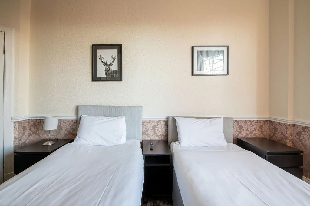 Two single beds with white bedding and pillows, bedside tables, a lamp on the left, a remote on the right, and framed pictures of a deer and a window on the wall above the beds.