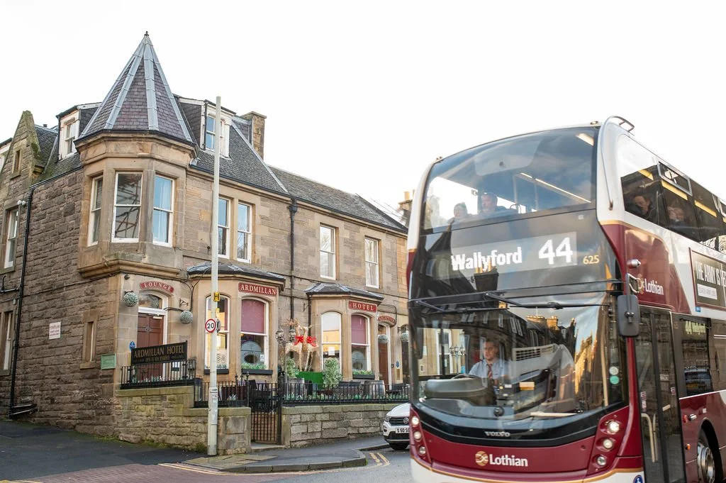 A historic stone building with turrets and windows, labeled as Ardillan Hotel, is situated on a street corner. In front there is a red double-decker bus with the route number 44 heading to Wallyford.