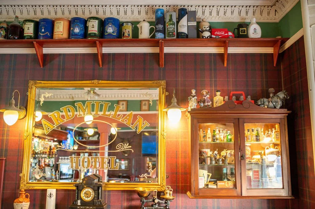 Interior of the Ardmillan Hotel with a large gold-framed mirror, decorative objects, a wooden display cabinet with glass doors containing bottles and items, and shelves with tins, mugs, and collectibles on the wall.