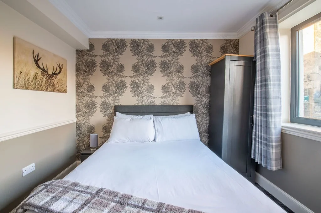 A bedroom with a double bed, white bedding, and pillows, a wall with floral wallpaper, a gray wardrobe next to a window with plaid curtains, and a landscape painting of a deer antler on the wall.
