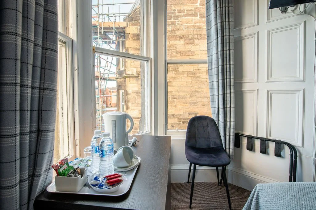 Interior of a room with a window, a small table with bottled water, a kettle, cups, and snacks, plus a black chair and a bed or couch in front of an ornate wall panel.
