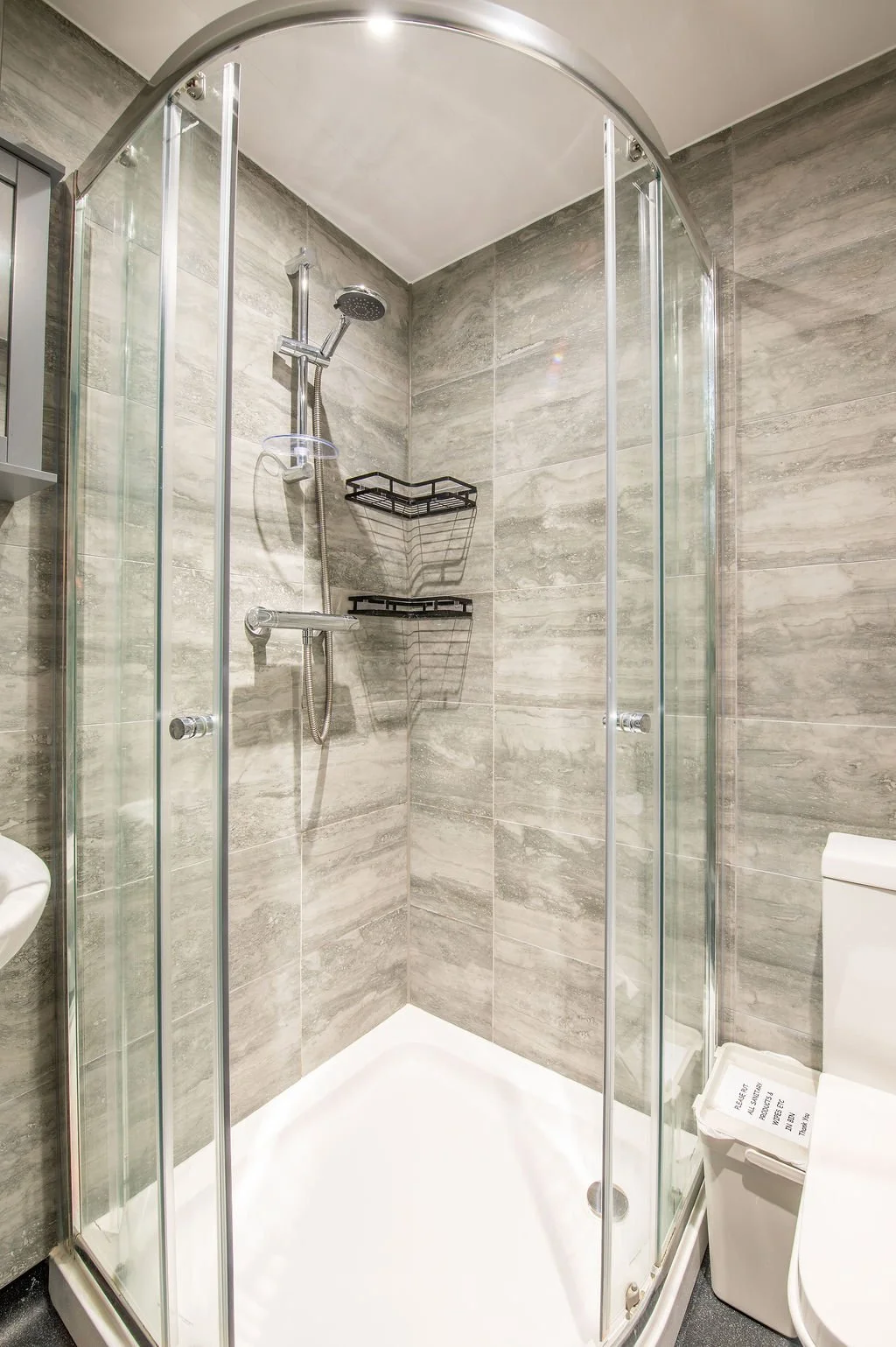 A modern bathroom shower with glass doors, gray stone tile walls, a chrome showerhead, a handheld shower wand, black wire shelves, and a white toilet nearby.