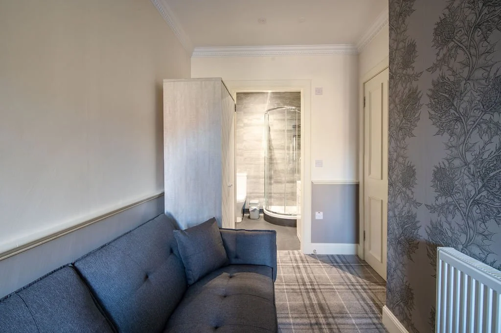 A living room with a gray sofa, a wardrobe, and a patterned wallpaper. In the background, a bathroom with a shower enclosure, toilet, and tiled flooring is visible.