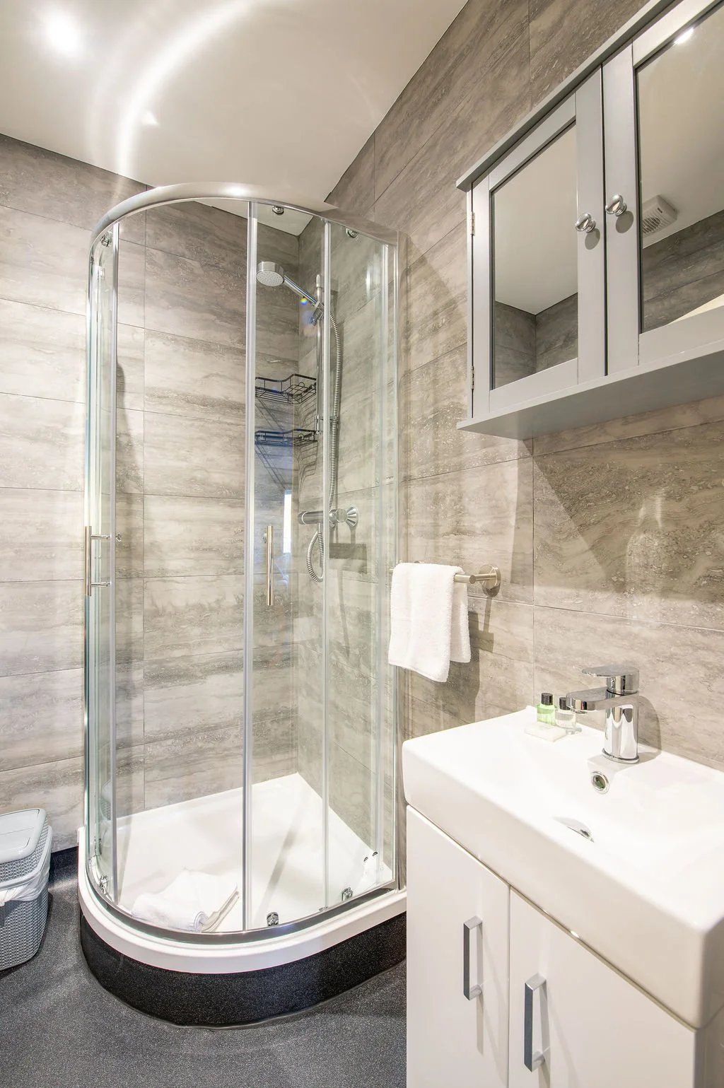 A modern bathroom with a corner shower enclosure, a small white sink cabinet with a mirror cabinet above, and a towel hanging on a wall-mounted towel bar.