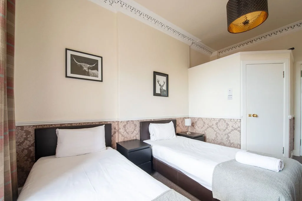 Hotel room with two single beds, side table, lamp, and framed animal artwork, in beige and brown decor.