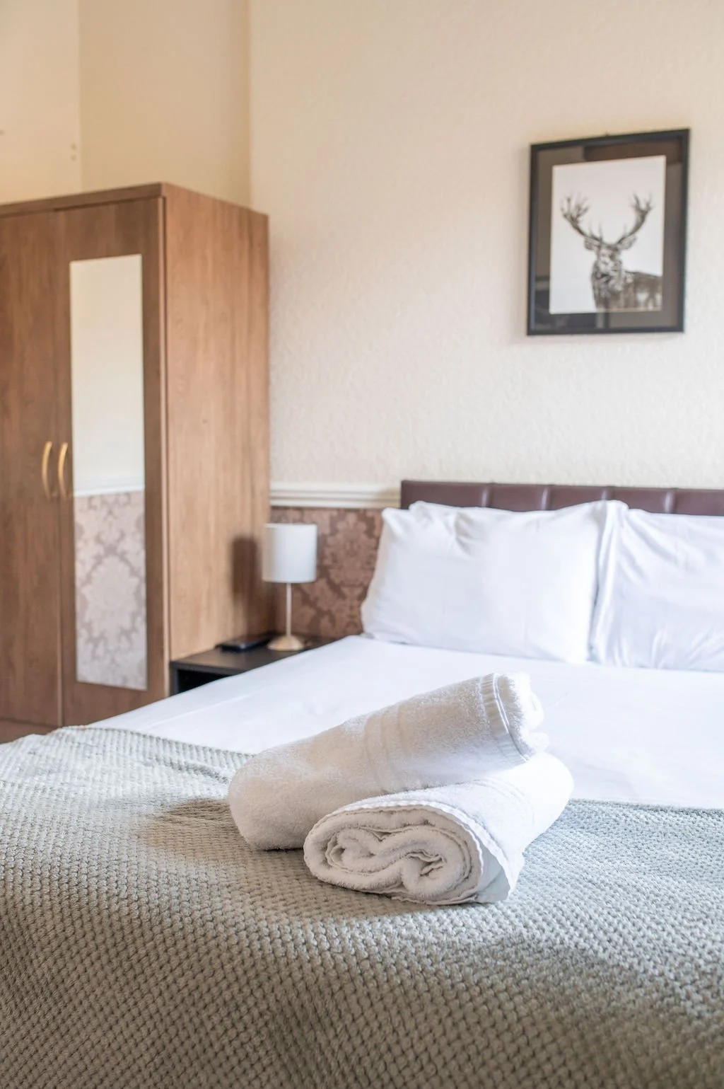 A neatly made bed with rolled white towels on a textured beige blanket in a hotel room. A wooden wardrobe, a small nightstand with a lamp, and a framed picture of a deer are visible in the background.