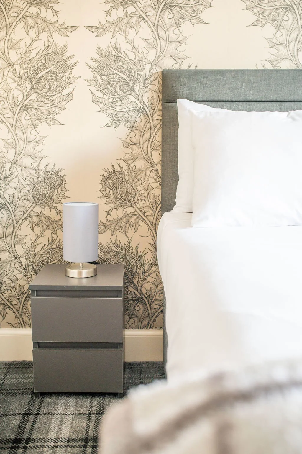 A bedside scene showing a nightstand with a lamp, a bed with white sheets and pillows, gray upholstered headboard, patterned wallpaper with artichoke illustrations, and a carpeted floor.