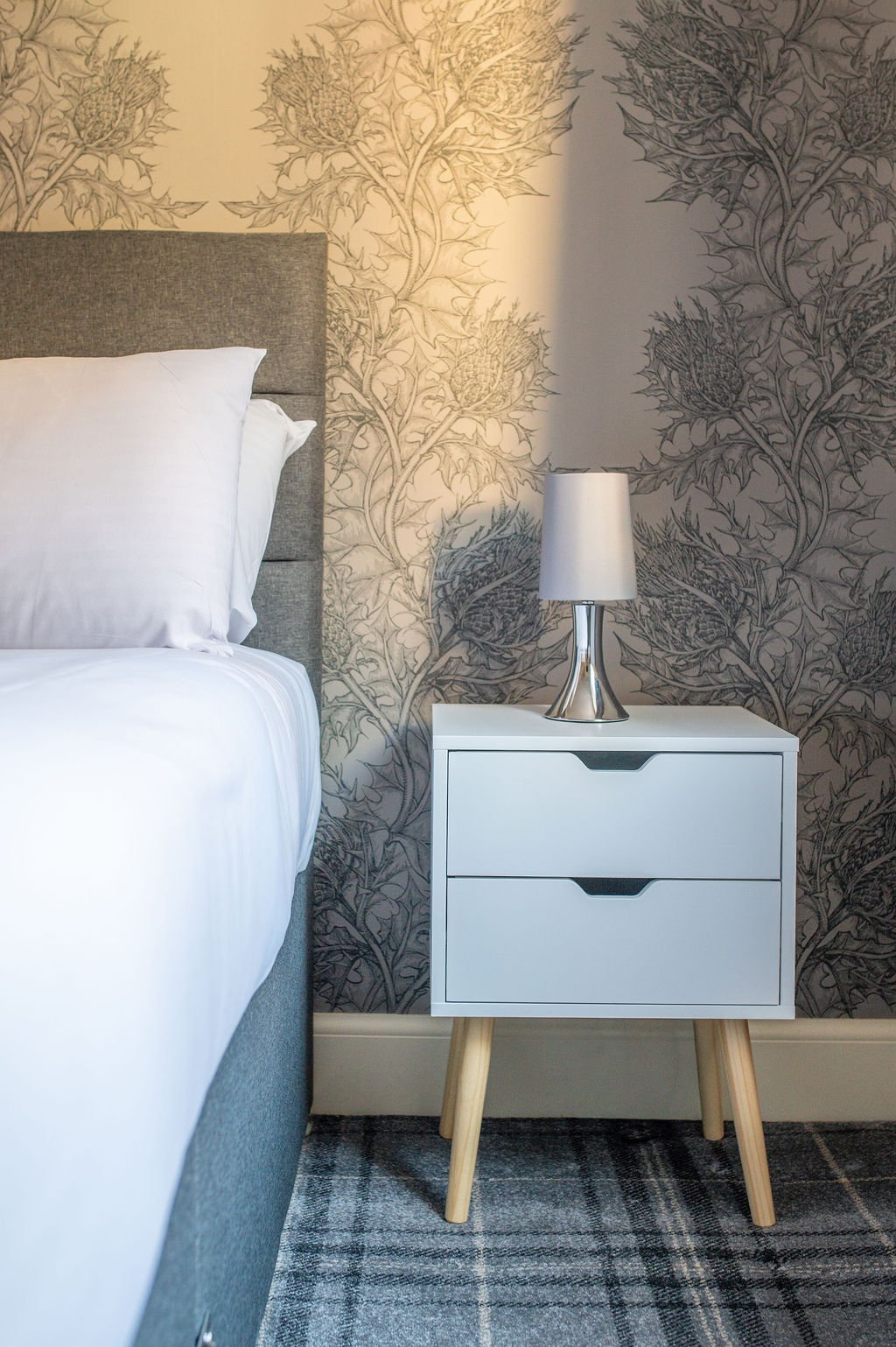 A bedroom nightstand with a white lamp, next to a bed with white bedding, against a large floral wallpaper with thistle-like plants and leaves, and a patterned rug on the floor.