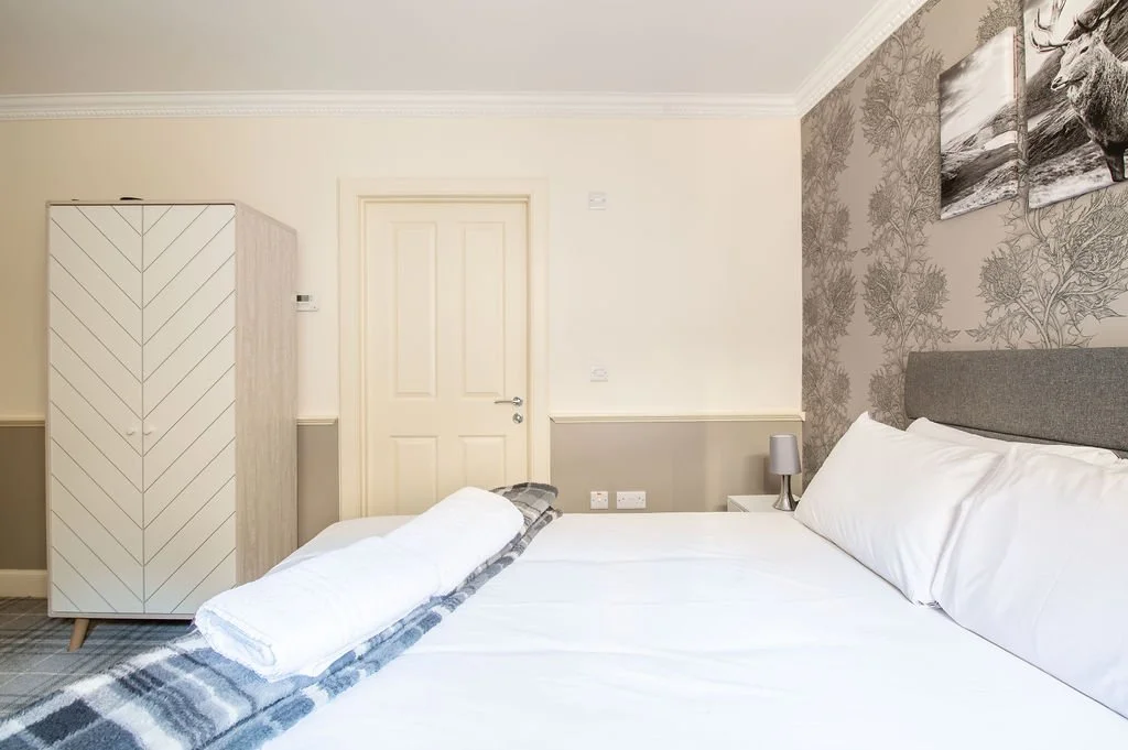 A neatly made bedroom with a white bed, gray headboard, two pillows, a bedside lamp, a white wardrobe, and a door. Artwork featuring a moose hangs on a patterned accent wall.
