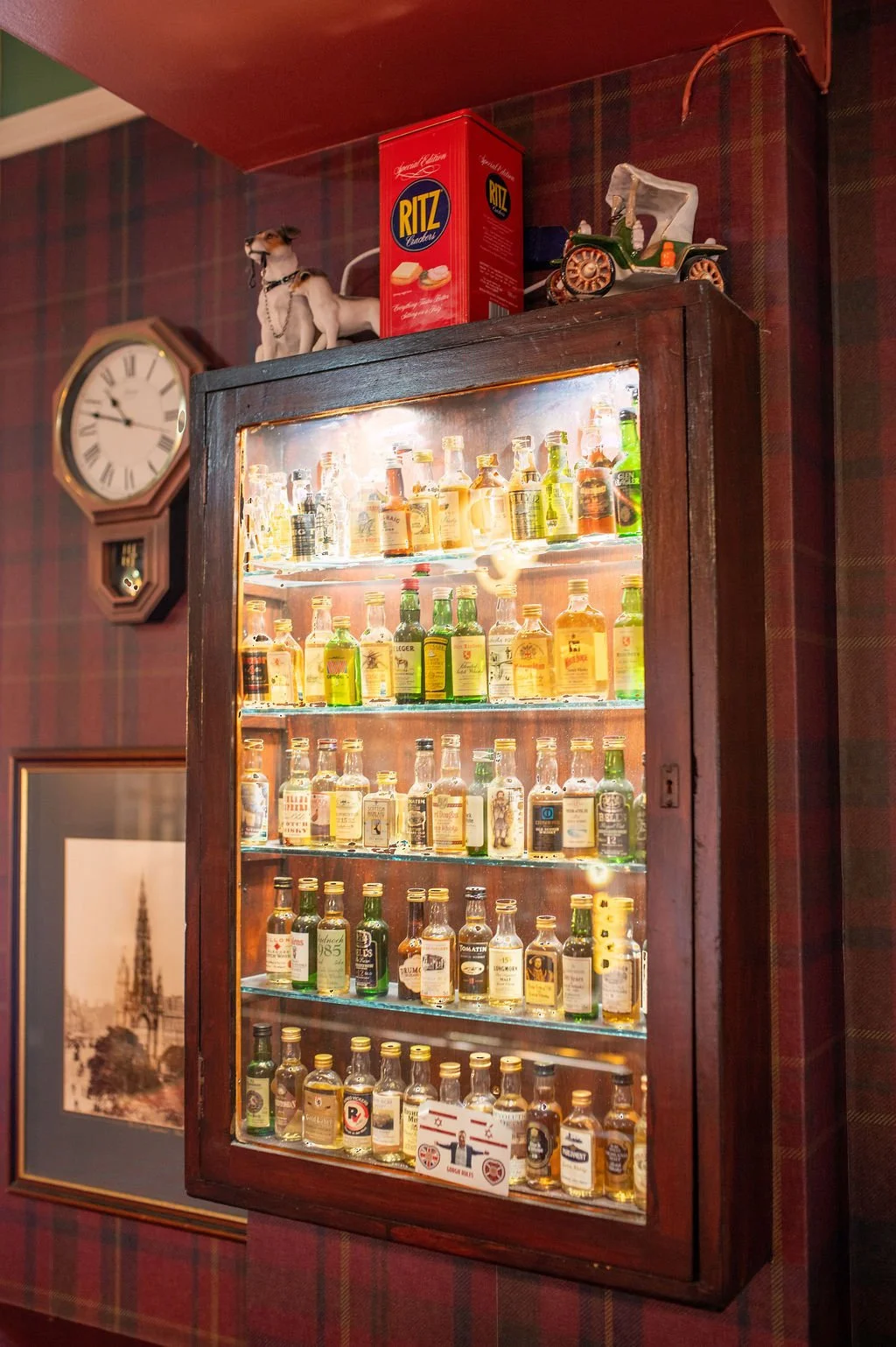 A vintage display cabinet filled with small collectible bottles, mounted on a wall with tartan wallpaper, featuring a clock and decorative items on top.