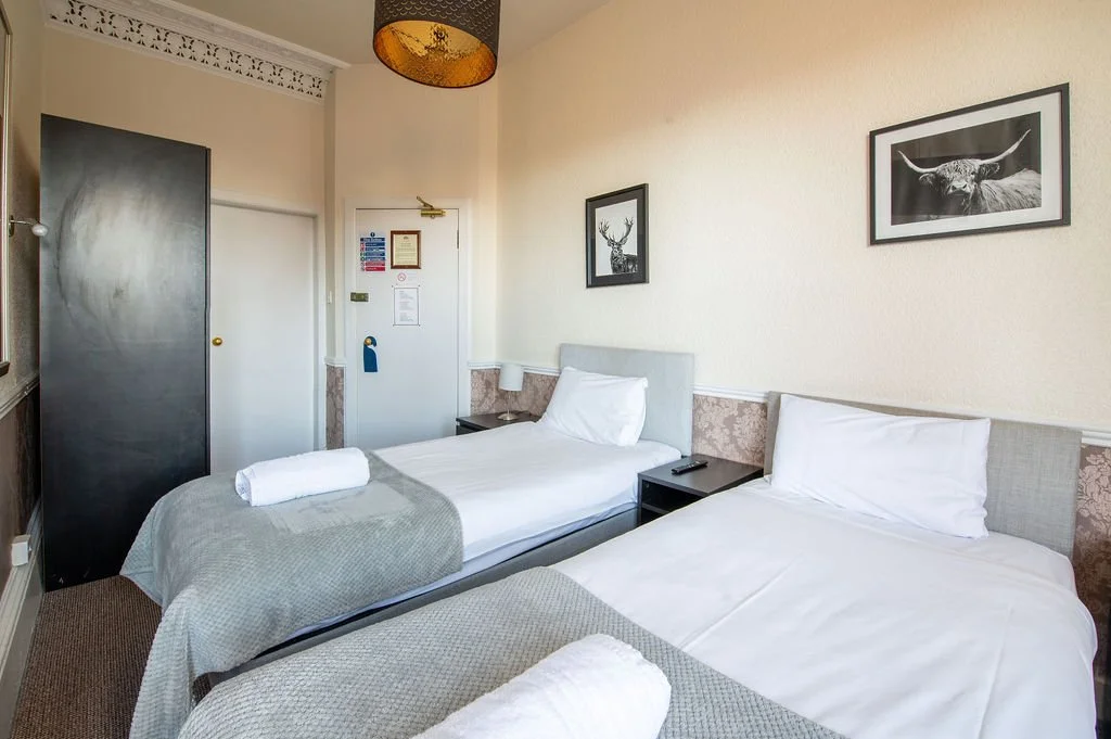 Two single beds in a hotel room with white linens, gray headboards, and rolled towels, with black and white artwork on the wall above, and a lamp hanging from the ceiling.