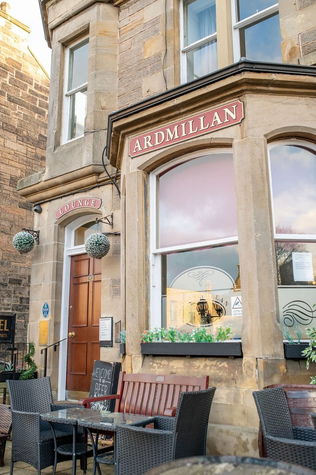 Exterior of a building with a sign reading 'Ardmillan' above a large window, and another sign reading 'Lounge' above the door. There are outdoor seating arrangements with chairs and tables, a small chalkboard sign welcoming guests with 'Welcome to ou
