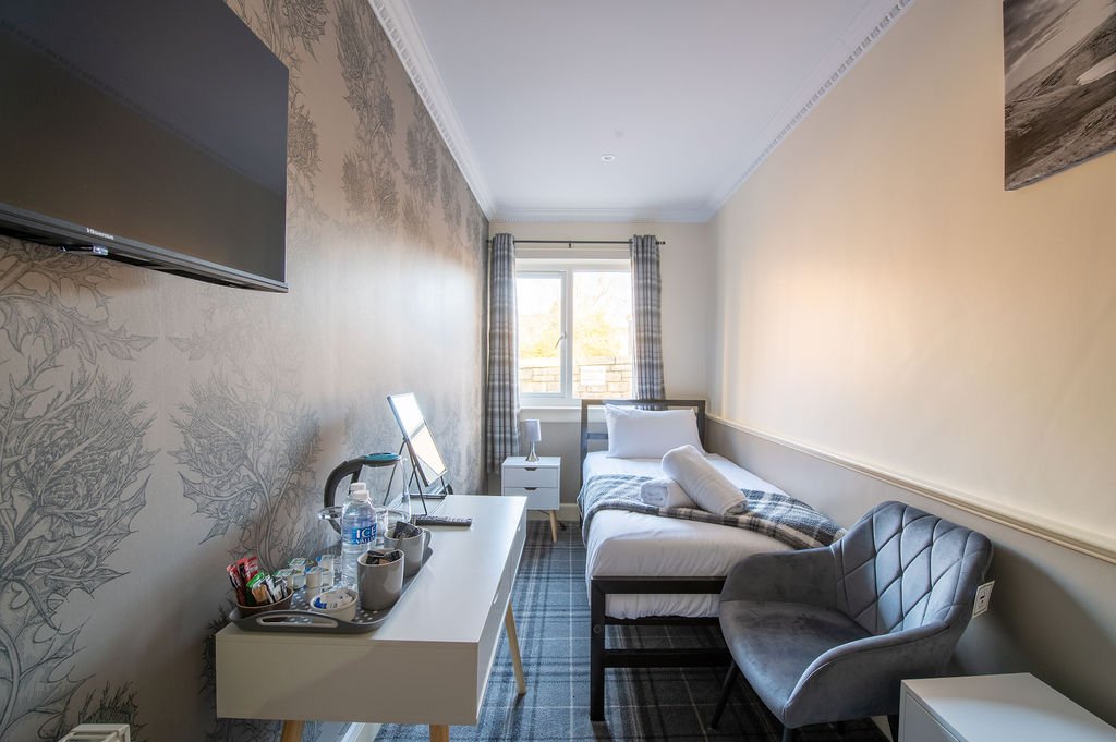 A small bedroom with a bed near the window, a gray armchair, a white desk with items on it, and a wall-mounted TV.