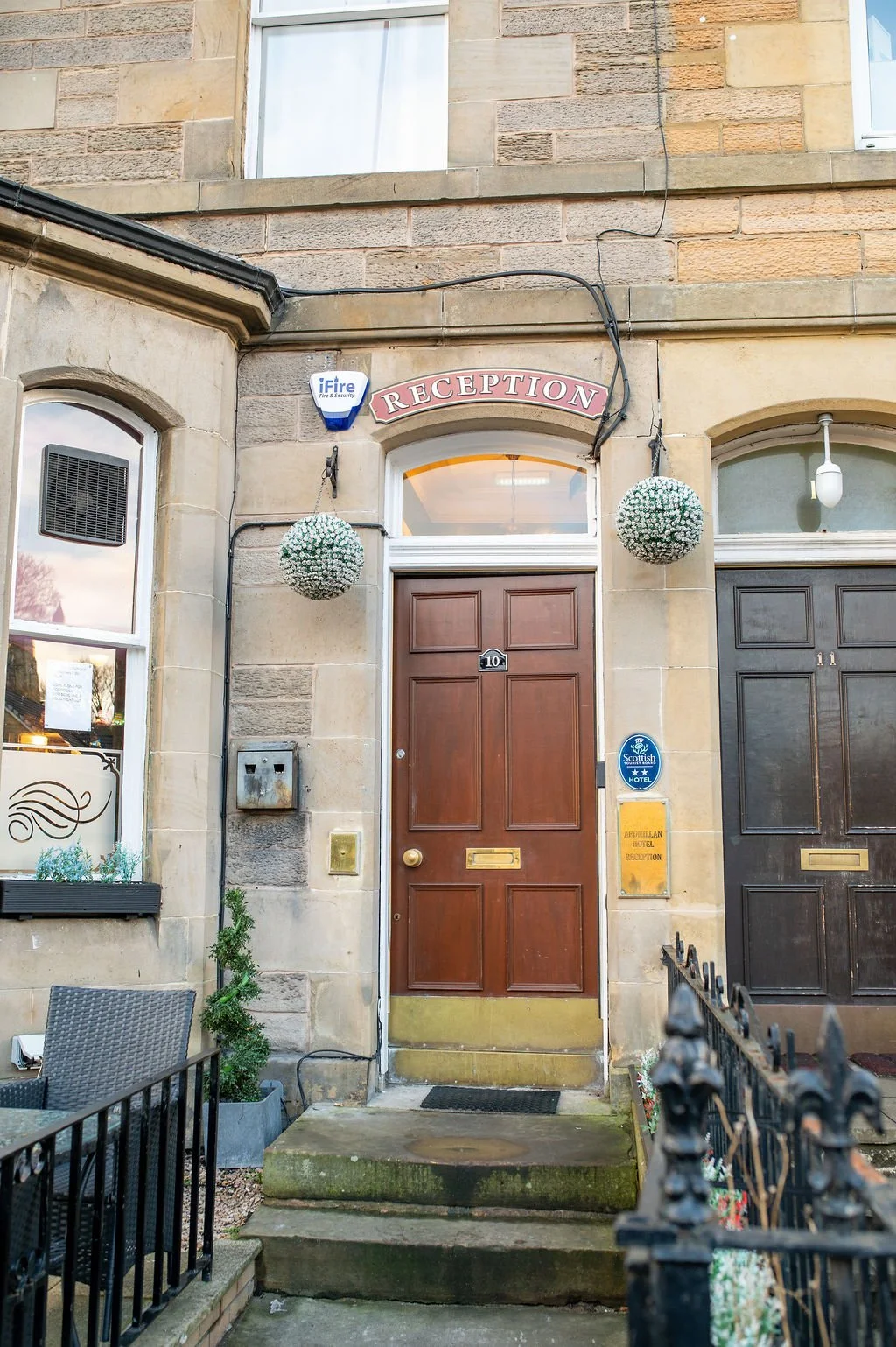 Wooden front door with a brass mail slot and door handle, above three concrete steps with moss, flanked by a black wrought iron fence, with hanging white flower arrangements and a 'Reception' sign above, part of a stone building with large windows.