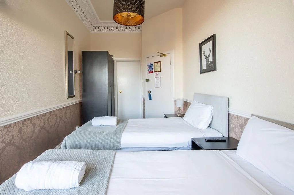 Hotel room with two beds, each with white linens and towels, a dark wardrobe, a bedside table with a lamp, a mirror on the wall, and a framed picture of a deer.