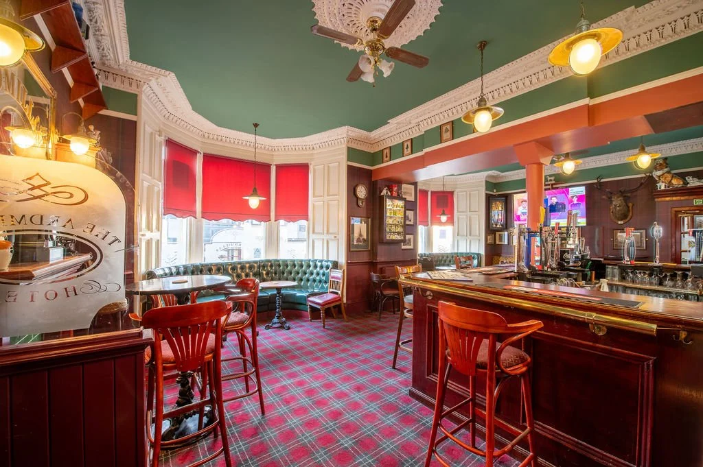 Interior of a vintage pub or bar with red and green decor, wood paneling, a curved bar counter with barstools, green upholstered seating, red curtains, and a patterned carpet.