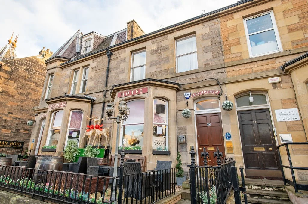 Exterior of a hotel with a reception entrance, decorated with holiday ornaments including reindeer and hanging baubles, and outdoor seating area with chairs and plants.