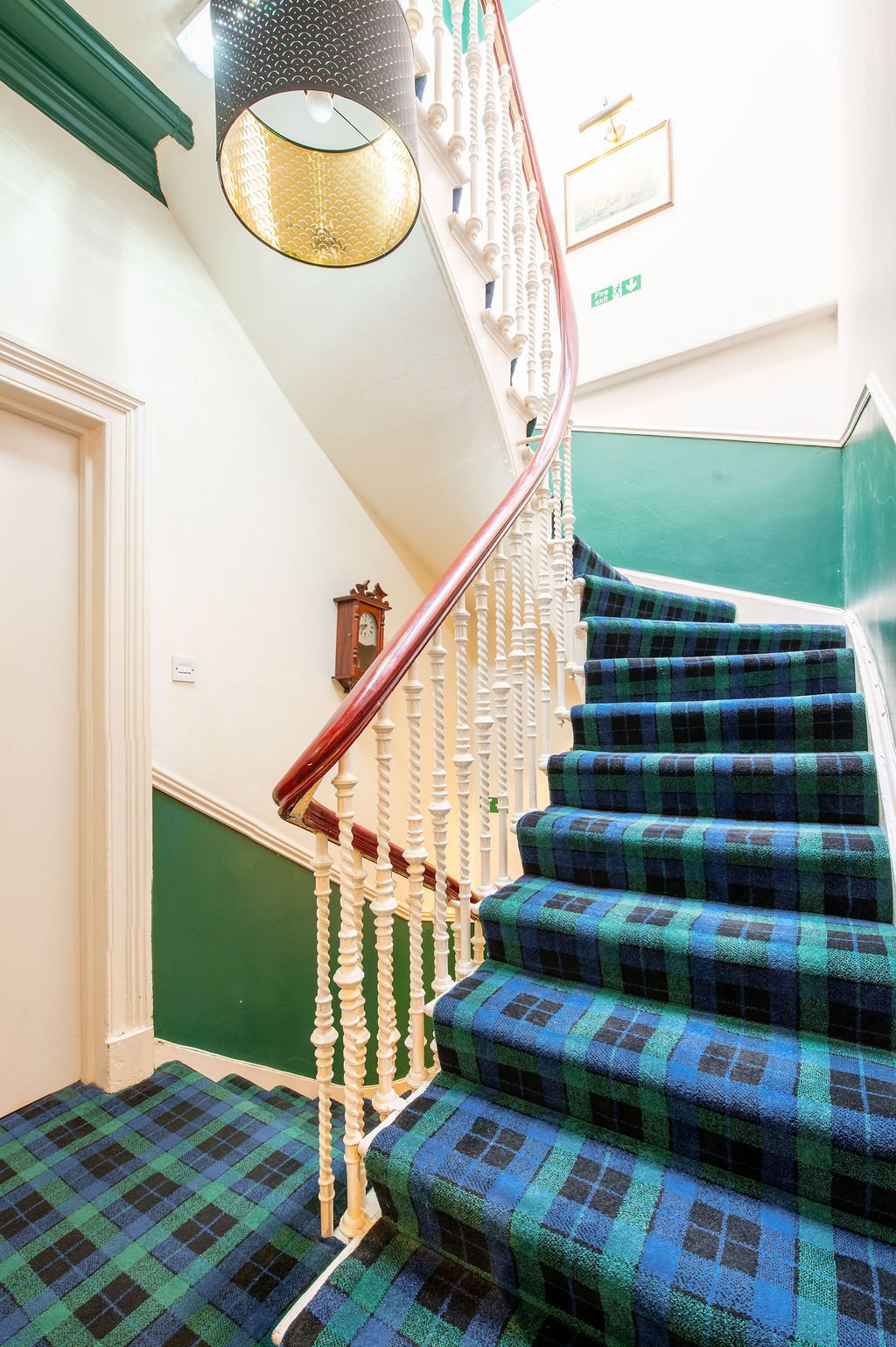 Spiral staircase with patterned blue, green, and black carpet, wooden handrail, white balusters, and green wall paneling. A vintage wall clock and ceiling light fixture are visible, with framed artwork on the white wall near the top of the staircase.