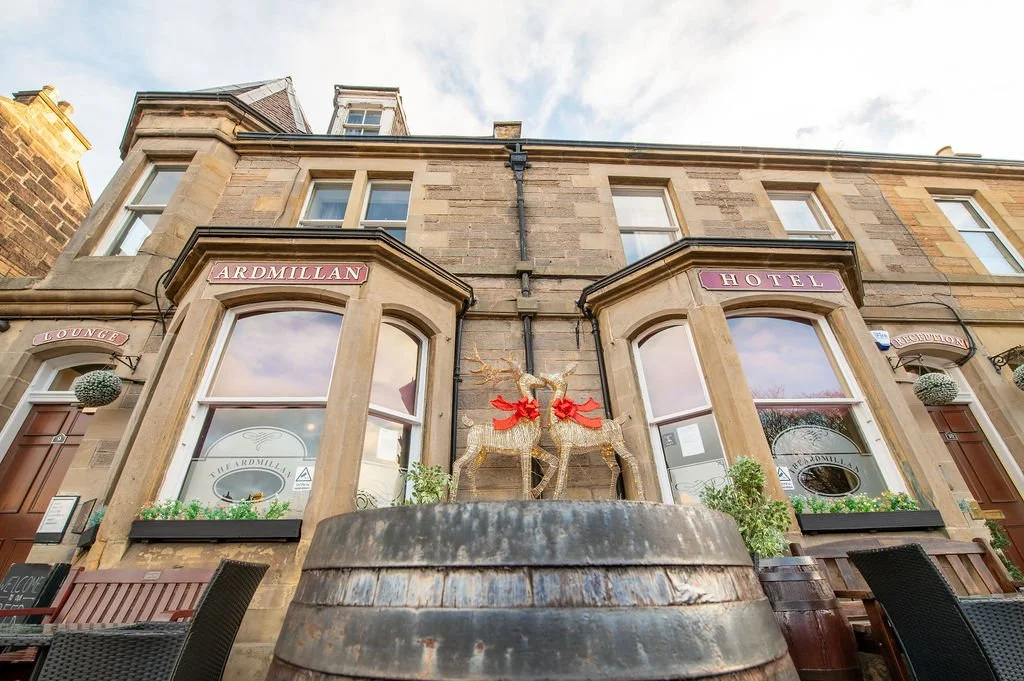 The exterior of a historic hotel with festive holiday decorations, including two gold reindeer figures with red bows, in front of large windows. The hotel has signs displaying the name "Ardmillan" and mentions "Lounge," "Reception," and "Hotel."