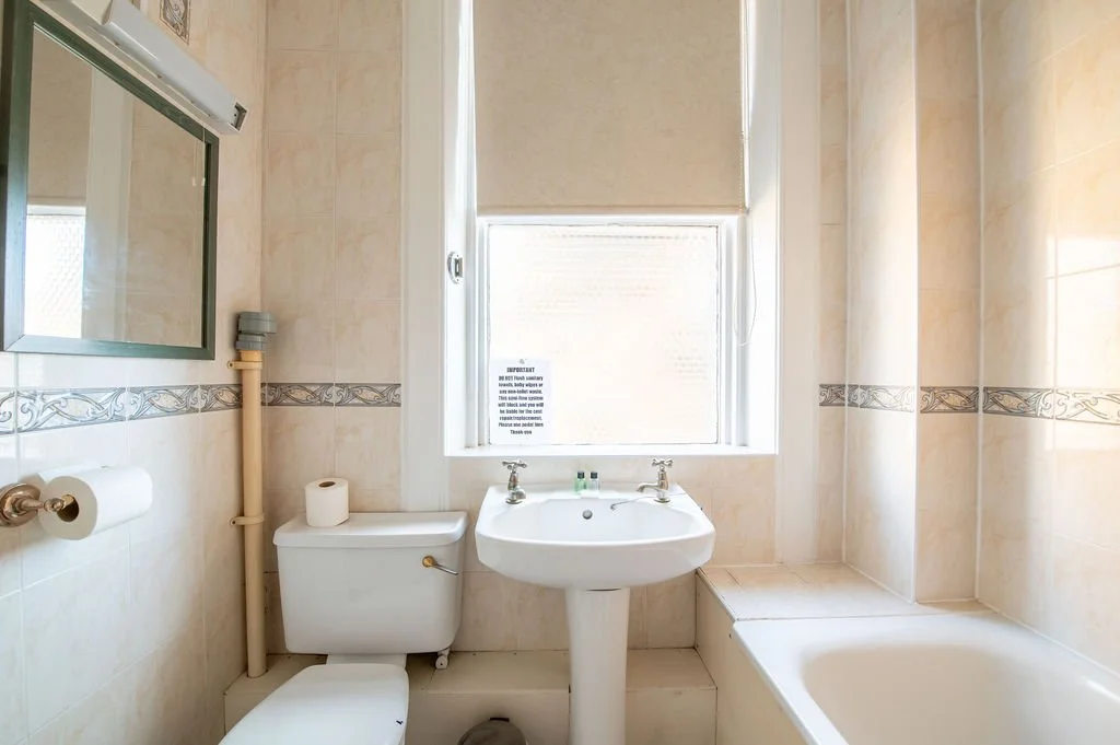 A small bathroom with beige tiled walls, a toilet with a roll of toilet paper on top, a pedestal sink with two soap bottles, a mirror, and a bathtub is seen. A window with a sign and a partially lowered beige blind is above the sink.