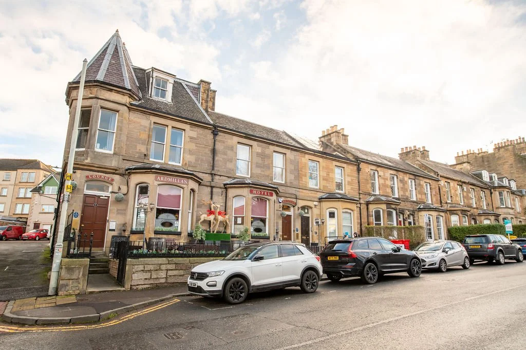 A row of historic stone buildings with a hotel and lounge, cars parked along the street, and a cloudy sky.