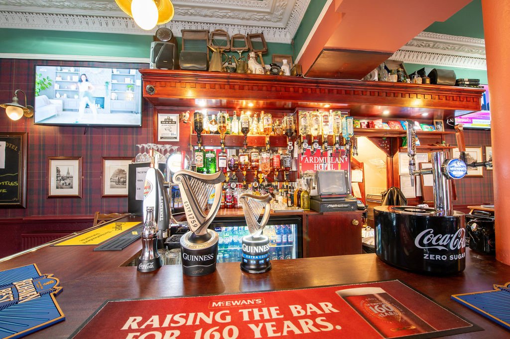 The bar features a wooden counter with beer taps, including Guinness, and a Coca-Cola Zero Sugar dispenser. Shelves behind the bar are stocked with various bottles of alcohol. There is a television mounted on the wall displaying an image of a person 