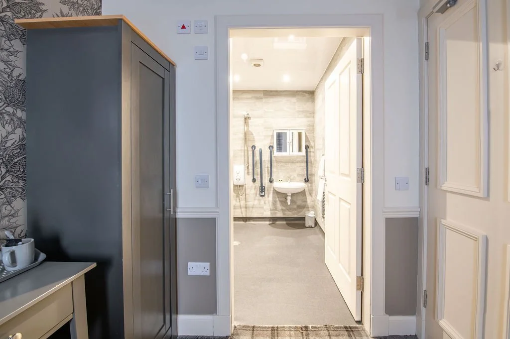 View into a clean, well-lit bathroom with gray tiled walls, a small sink, and accessible grab bars.
