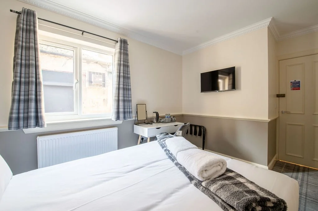 A hotel room with a bed, a window with plaid curtains, a wall-mounted TV, a small desk with a mirror, a chair, and a towel and blanket on the bed.