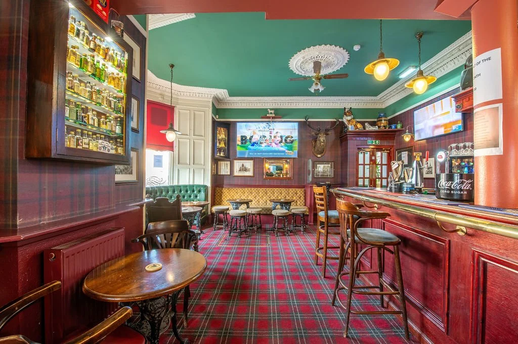 Interior of a pub with green and red walls, wooden furniture, a tartan carpet, a bar with stools, and a television.