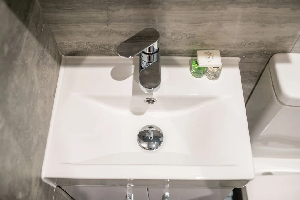 White bathroom sink with a silver faucet, two small bottles, and a white soap dish on the side, next to a toilet in a bathroom with gray tiled walls.