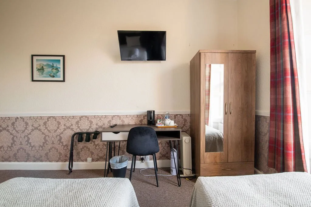 A hotel room with two beds, a small desk and chair, a wardrobe with mirror, a wall-mounted TV, and a window with red plaid curtains.