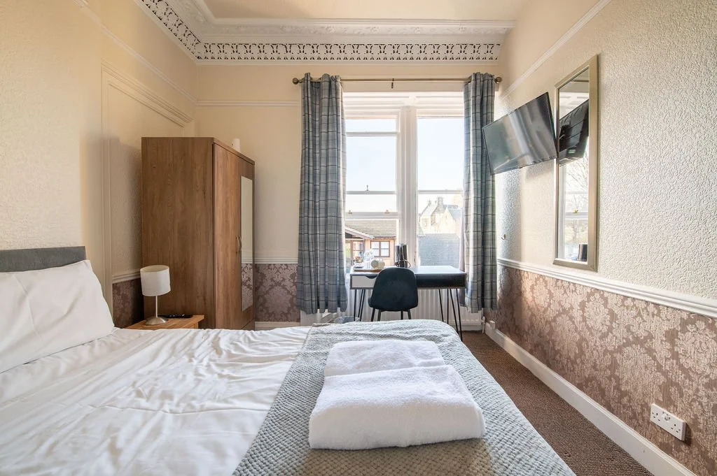 A cozy hotel room with a bed, bedside lamp, wardrobe, desk with chair, and wall-mounted TV, featuring large window with blue curtains allowing sunlight.