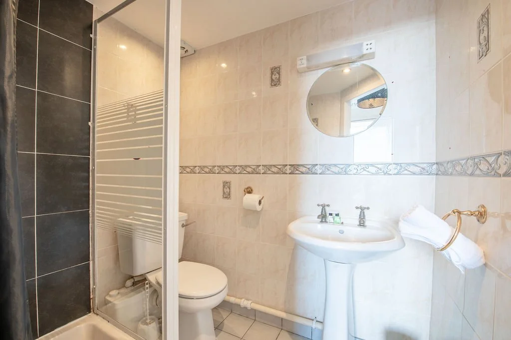 A small, clean bathroom with a shower, toilet, and pedestal sink. The walls are tiled in beige with decorative accents, and there is a round mirror above the sink. A towel is hanging on a gold towel ring beside the sink. The shower enclosure has a sl