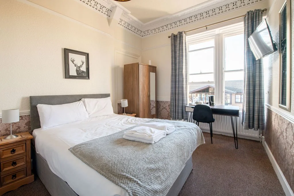 A bedroom with a large bed, nightstands, lamps, a wardrobe, a desk with a chair, a window with curtains, and a wall-mounted TV.