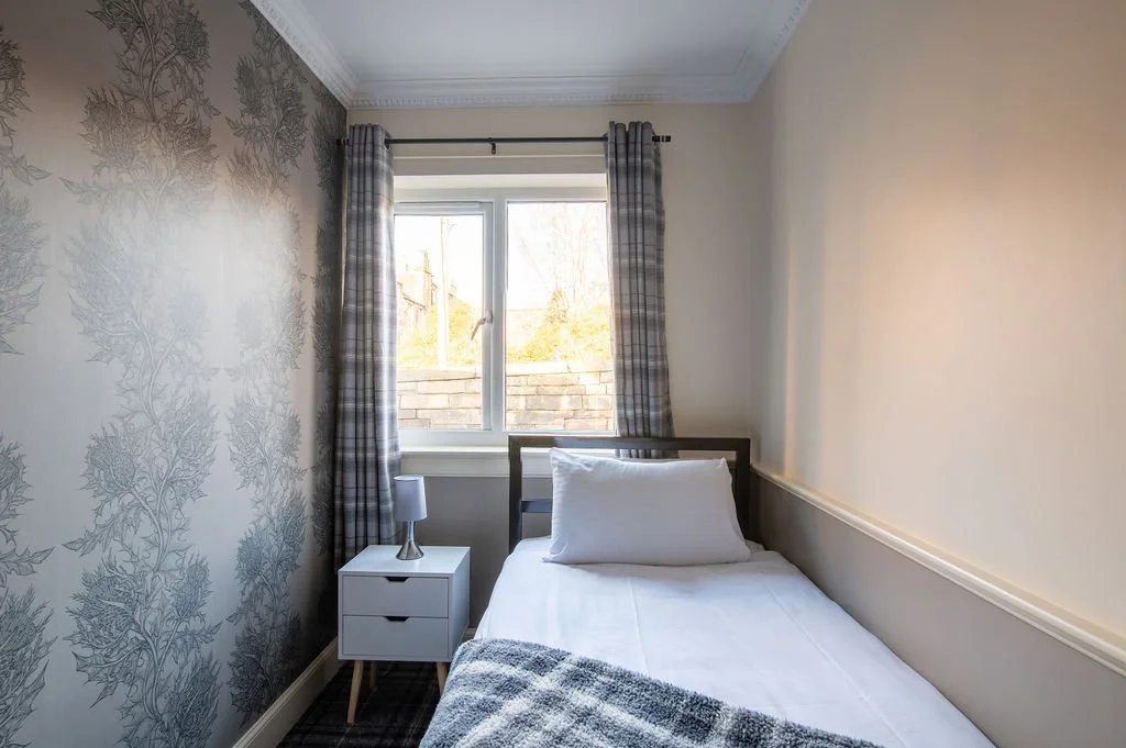 A small bedroom with a single bed, white bedding, a pillow, plaid curtains, a window with a view of trees and a brick wall outside, and a white nightstand with a small lamp.