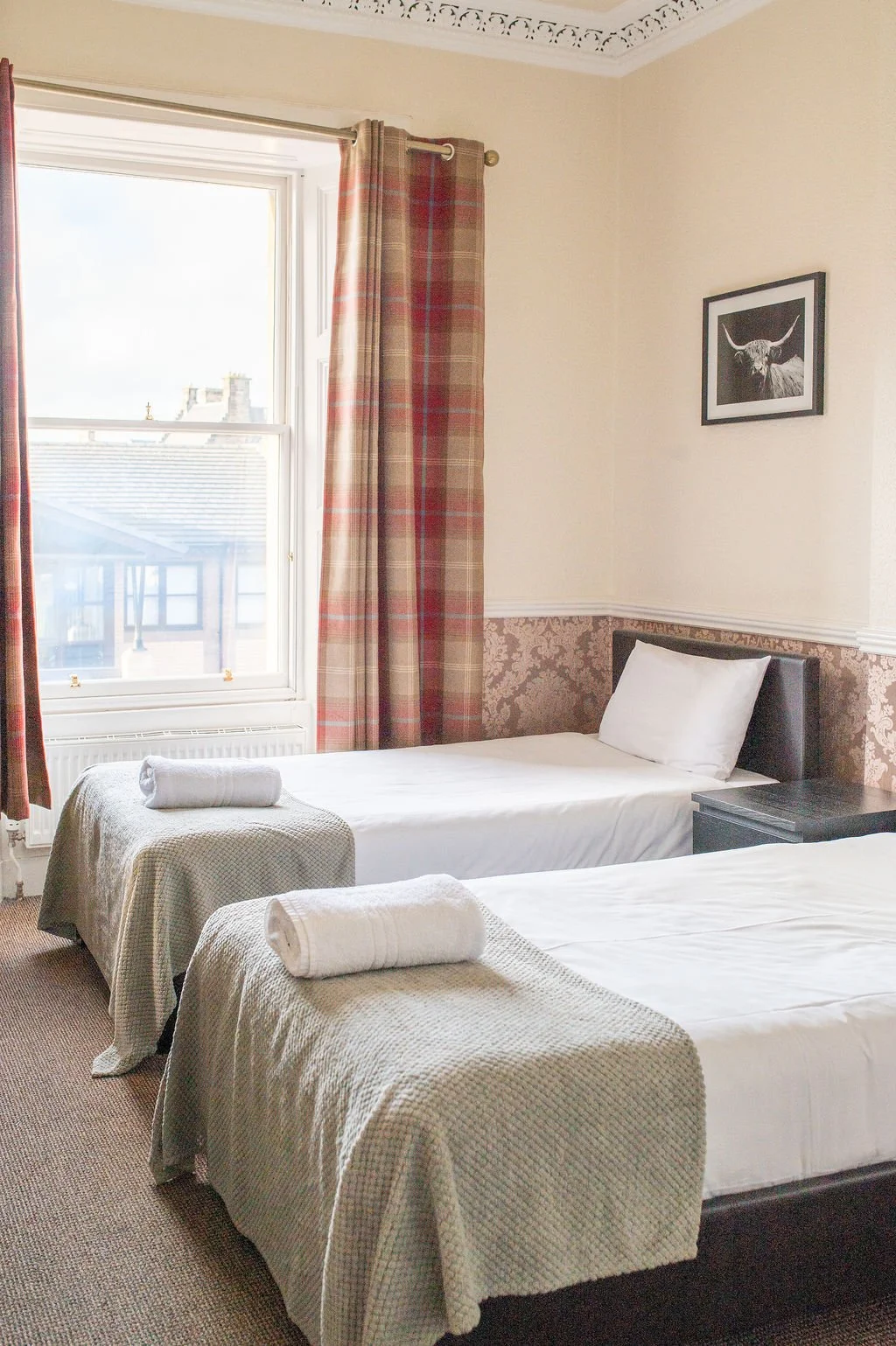 A bedroom with two single beds, each with a rolled towel, beige bedspreads, and white pillows. A window with plaid curtains and a picture of a highland cow on the wall.