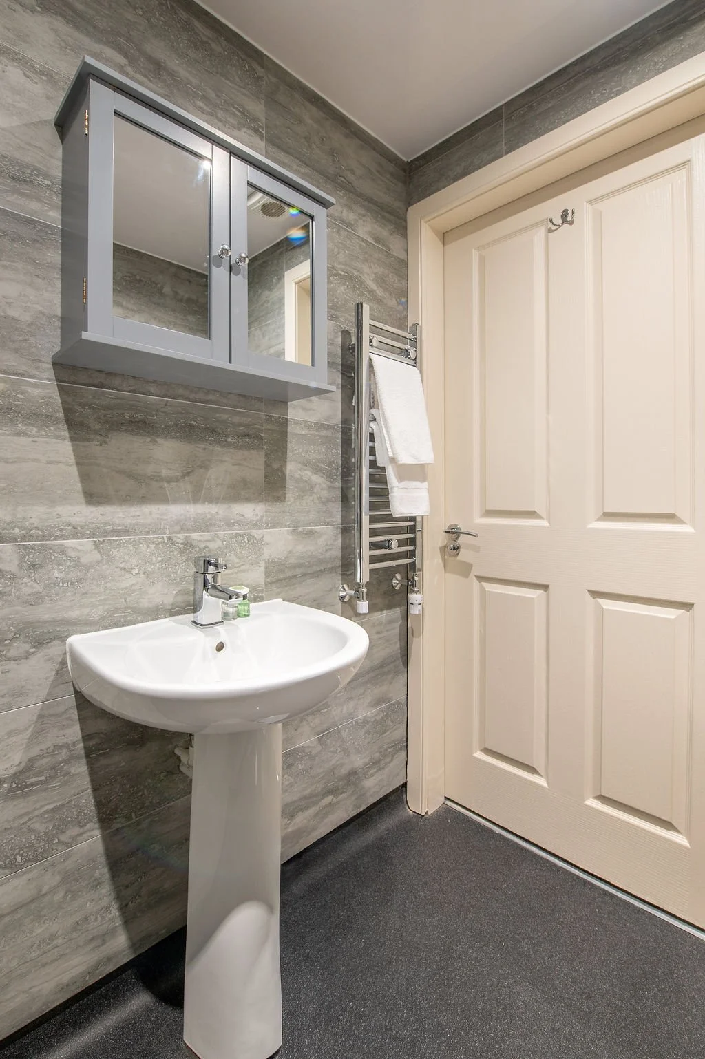 A small bathroom with gray tiled walls, a white pedestal sink, a gray wall-mounted cabinet with mirrors, a towel rack with white towels, and a cream-colored door.