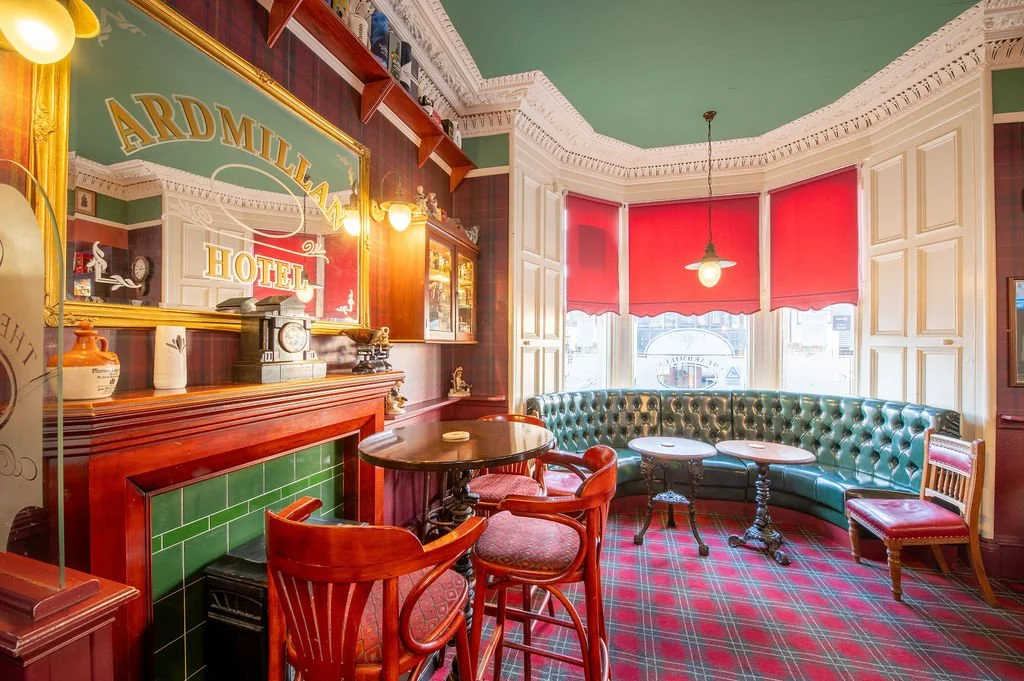 Interior of a vintage hotel lounge with green tufted curved sofa, red window shades, and wooden furnishings, reflecting an old-fashioned, cozy atmosphere.