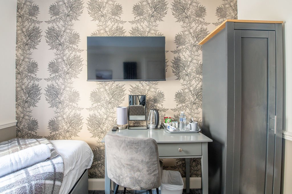 A hotel room with a bed, a wall-mounted TV, a desk with a chair, a mirror, a coffee machine, a kettle, water bottles, and a wardrobe. The wall has a patterned wallpaper with large floral designs.