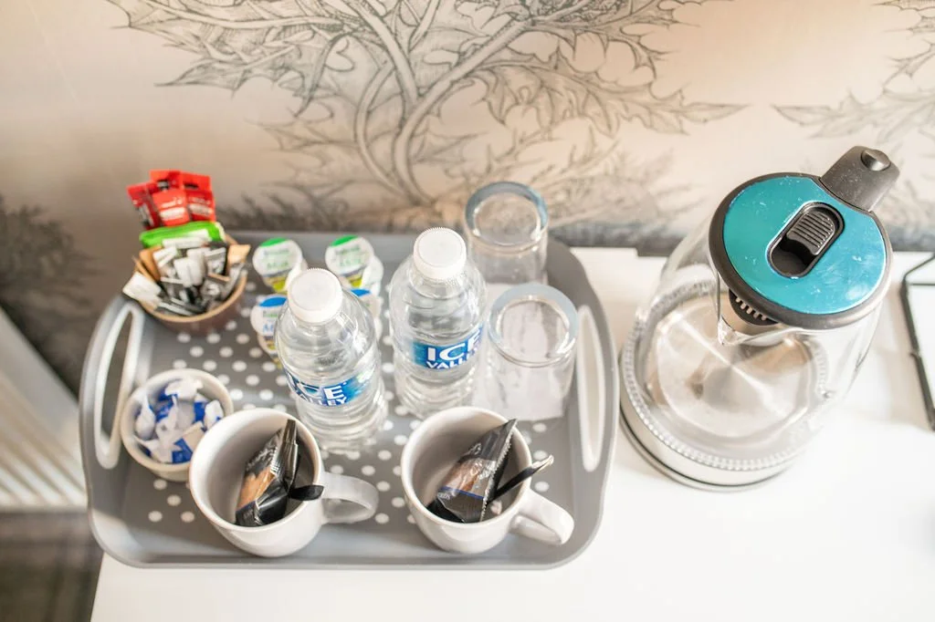 Tray of bottled water, coffee packets, sugar, and creamer cups beside a glass water pitcher with a water filter on a table.