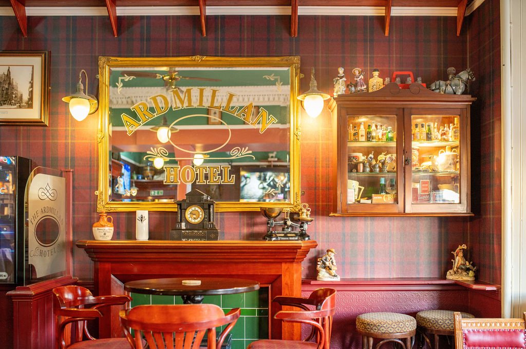 Interior of the Ardmillan Hotel with wood-paneled walls, vintage decor, a mirror with the hotel name, a fireplace with a green tiled base, wooden chairs, a glass display cabinet filled with collectibles, and small figurines on the shelves.