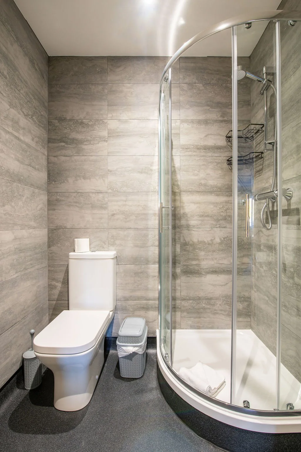A modern bathroom with a toilet, a trash bin, and a glass shower enclosure with a white towel inside.
