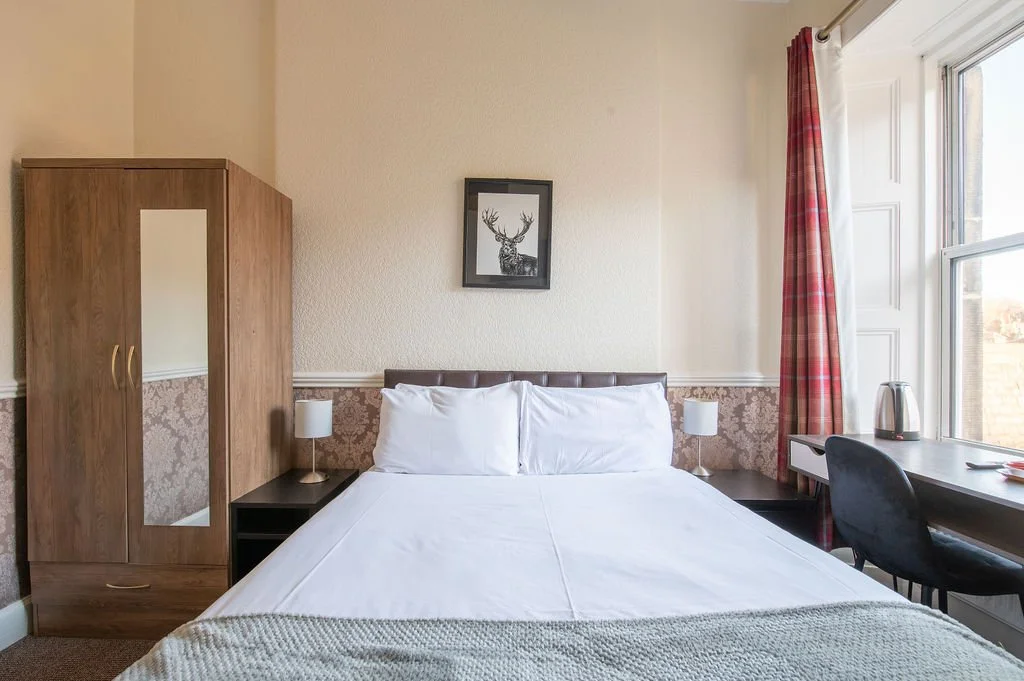 A neatly made bed with white pillows and bedding, flanked by two small lamps, in a cozy hotel room with a wooden wardrobe, side table, desk, chair, and a window with red checkered curtains.