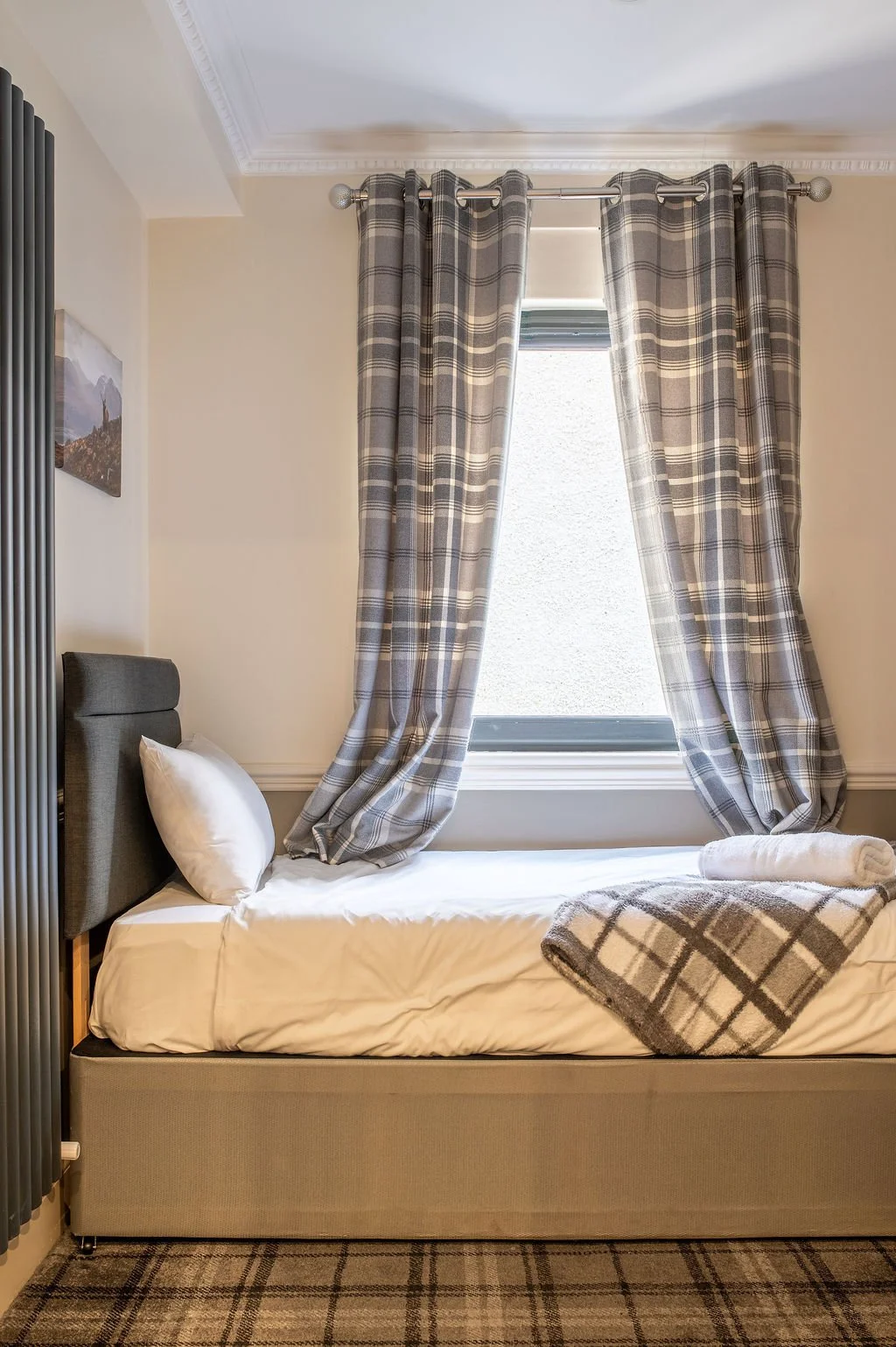 A single bed with a gray headboard and white bedding, positioned next to a window with plaid curtains. A rolled towel and a plaid blanket are on the bed, with matching plaid curtains hanging in front of the window. The room has beige walls with crown
