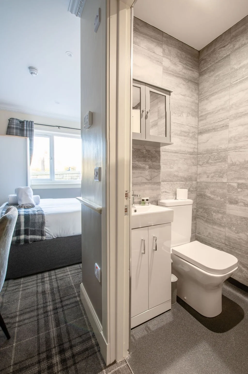 Interior of a small bathroom with grey tiled walls, a white toilet, a white sink with a cabinet, and a wall-mounted cabinet with double glass doors.