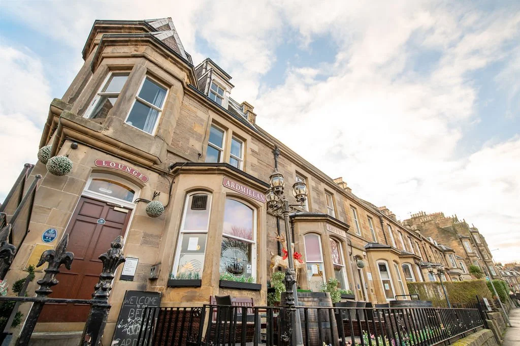 Historic stone building with bay windows, a sign for 'Lounge Ardmillan', and decorative street lamps, set against a partly cloudy sky.