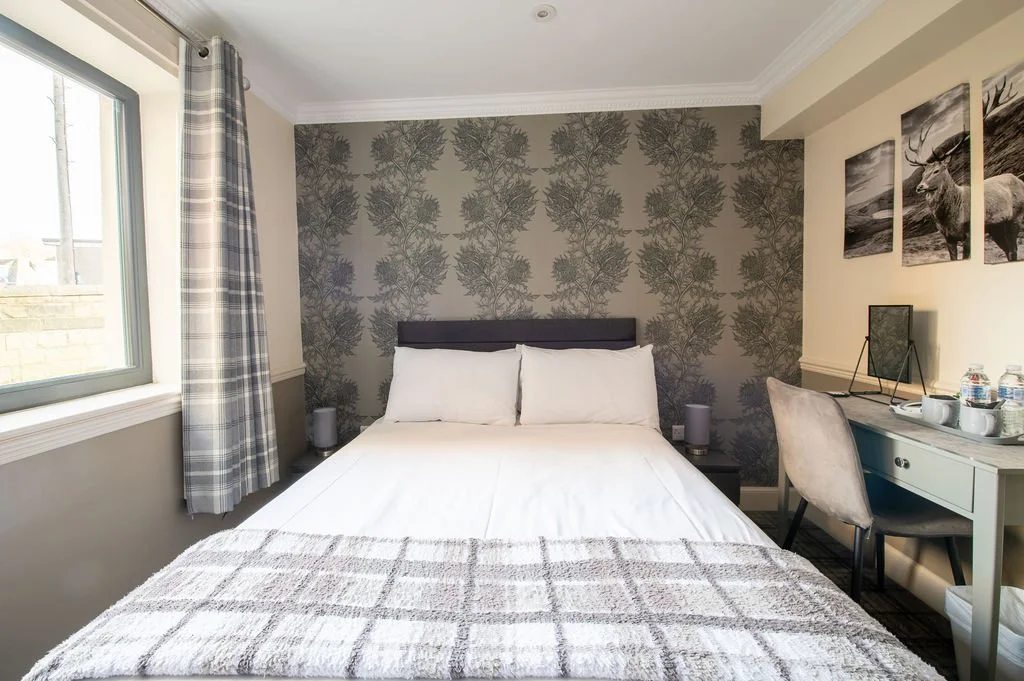 A bedroom with a bed against a patterned wallpapered wall, featuring white bedding and pillows. To the left, there's a window with plaid curtains. On the right side, there is a desk with a chair, art supplies, and several water bottles. There are bla